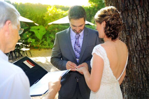 A groom in a suit places a ring on the brides finger during an outdoor wedding ceremony. The bride wears a white lace dress with an open back, and an officiant holds a folder nearby. Sunlight filters through the trees.