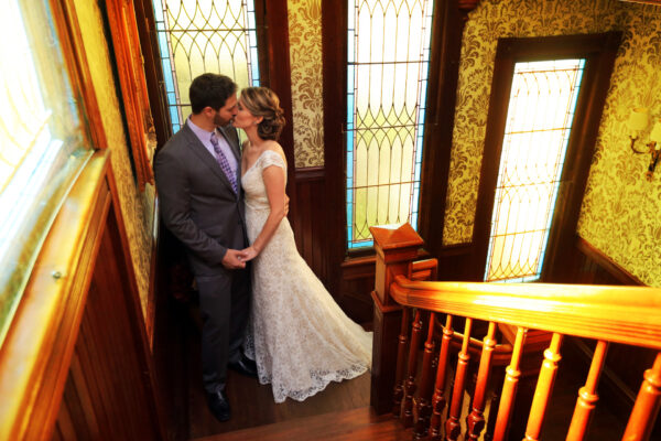 A bride and groom stand on a wooden staircase in a vintage house, holding hands and kissing. The bride wears a white lace dress; the groom is dressed in a gray suit. Stained glass windows and patterned wallpaper surround them.