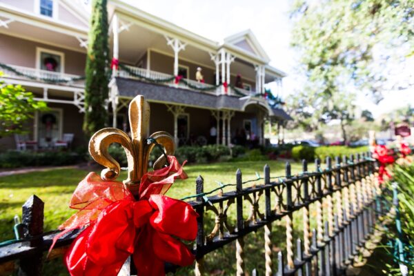 A close-up of a black iron fence with a gold fleur-de-lis finial and red ribbon, in front of a large Victorian-style house decorated with red bows on the porch and windows, surrounded by greenery.
