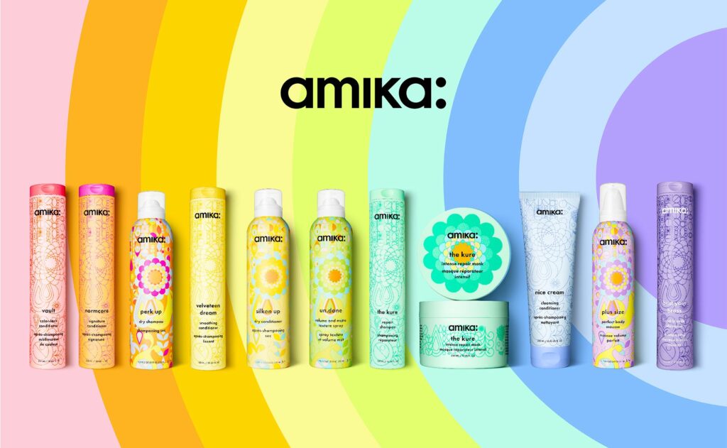 Colorful Amika hair care products in patterned bottles and jars are arranged in a row against a rainbow gradient background, with the black Amika logo above.