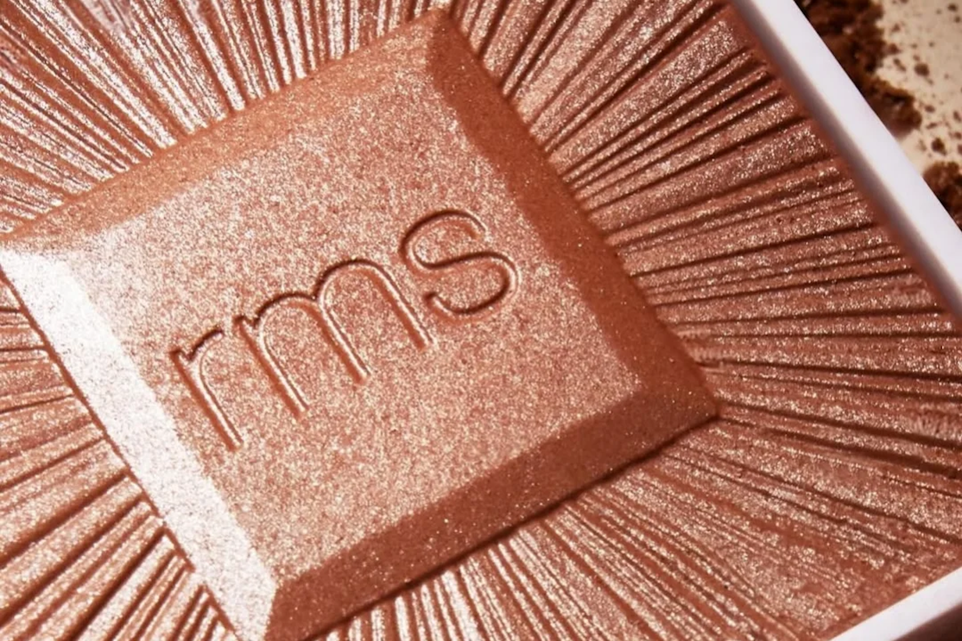 A close-up of a shimmery, rose-gold makeup highlighter with rms embossed in the center, featuring a radiant, sunburst design.
