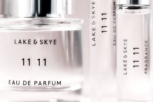Close-up of several glass perfume bottles labeled Lake & Skye 11 11 Eau de Parfum and Fragrance, with minimalist black text on clear bottles against a pale, softly blurred background.