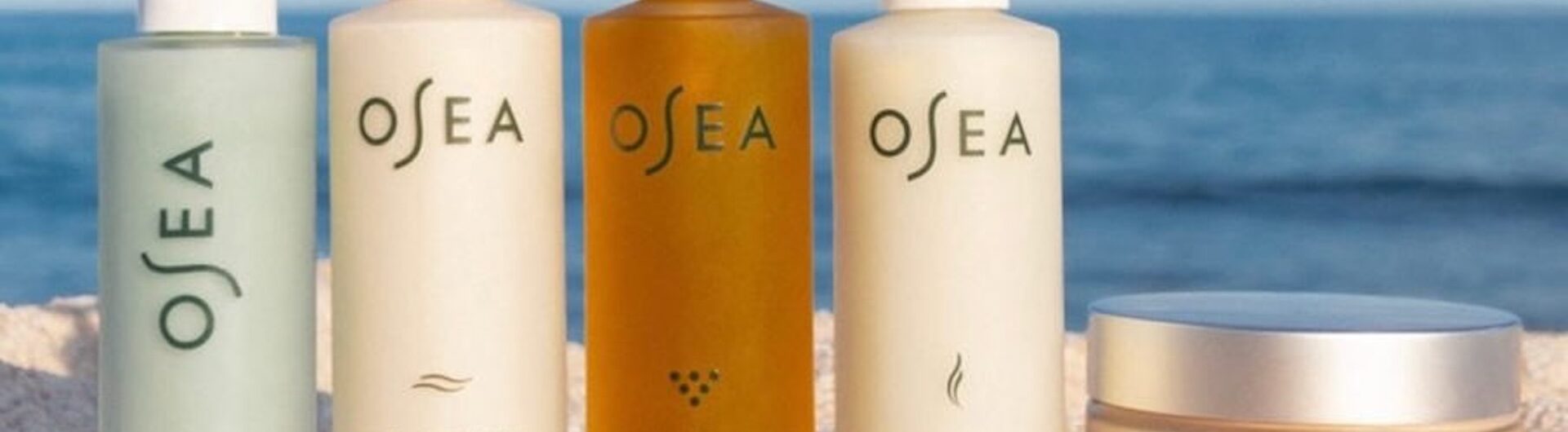 Five OSEA skincare products, including serums, lotions, and body butter, are arranged side by side on a sandy surface with a blue sky background.