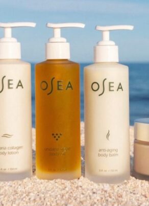Five OSEA skincare products, including serums, lotions, and body butter, are arranged side by side on a sandy surface with a blue sky background.