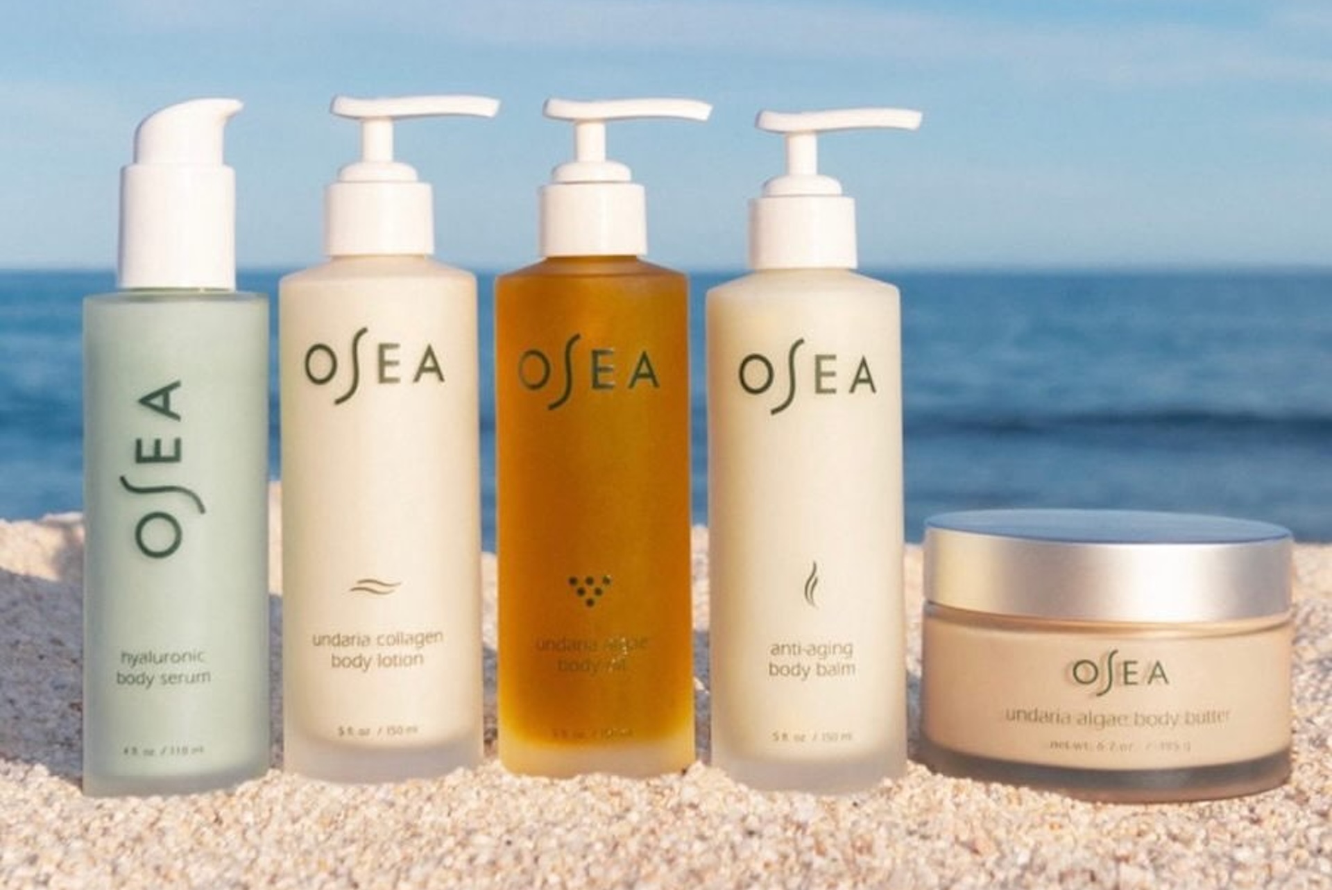 Five OSEA skincare products, including serums, lotions, and body butter, are arranged side by side on a sandy surface with a blue sky background.