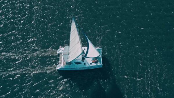 A white sailboat with raised sails glides across deep blue water, viewed from above. Sunlight reflects off the rippling surface, creating a textured backdrop around the boat.