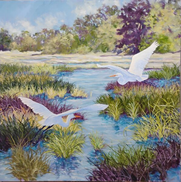 Two white egrets with outstretched wings are flying over a marsh with tall green grasses and shallow blue water, surrounded by distant trees under a partly cloudy sky.