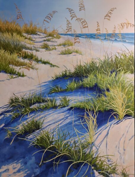 Sunlit sand dunes covered with green grasses and tall sea oats stretch toward a calm blue ocean under a clear sky. Shadows of the grasses create deep blue patterns on the sand.