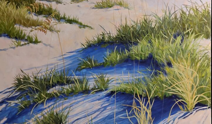 Sunlit sand dunes covered with green grasses and tall sea oats stretch toward a calm blue ocean under a clear sky. Shadows of the grasses create deep blue patterns on the sand.