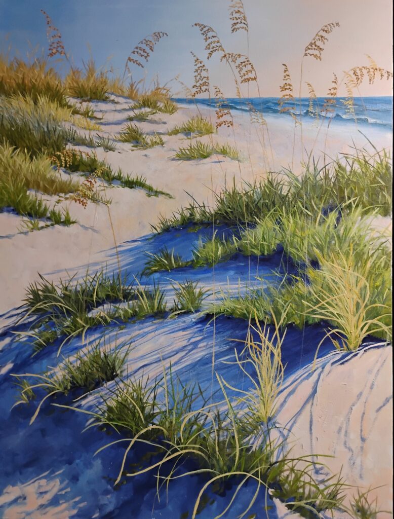 Sunlit sand dunes covered with green grasses and tall sea oats stretch toward a calm blue ocean under a clear sky. Shadows of the grasses create deep blue patterns on the sand.