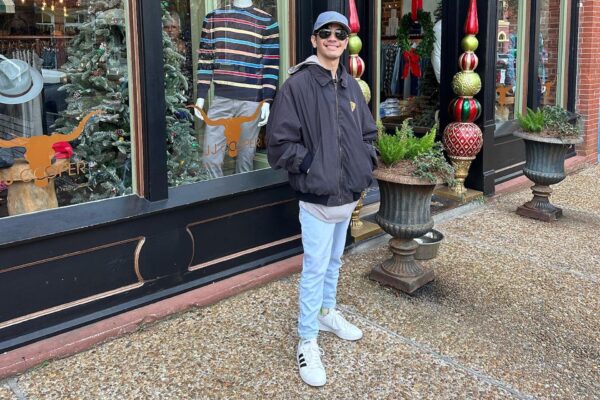 A person wearing a black jacket, blue jeans, white shoes, and sunglasses stands smiling on a sidewalk in front of a store decorated with Christmas trees and large ornaments.