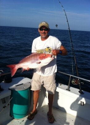 A man in sunglasses and a cap stands on a boat holding a large red snapper fish, with fishing rods and the ocean visible in the background under a clear sky.