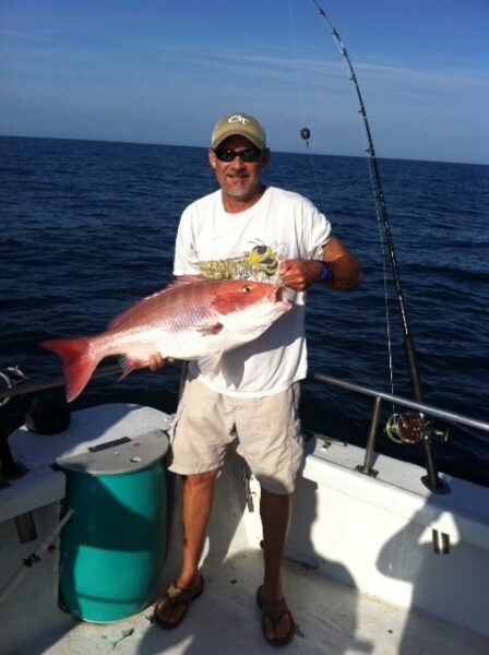 A man in sunglasses and a cap stands on a boat holding a large red snapper fish, with fishing rods and the ocean visible in the background under a clear sky.