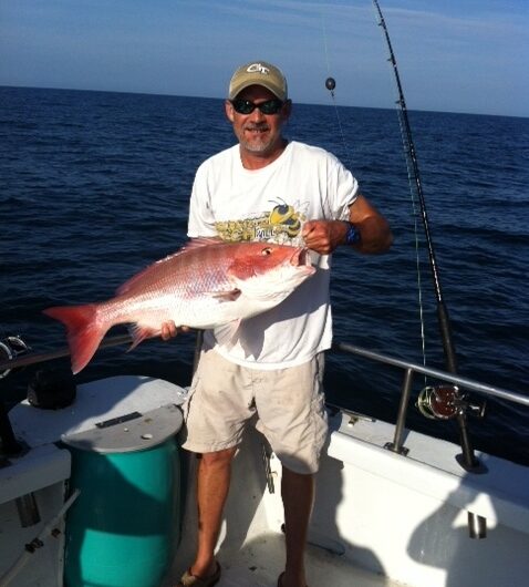 A man in sunglasses and a cap stands on a boat holding a large red snapper fish, with fishing rods and the ocean visible in the background under a clear sky.