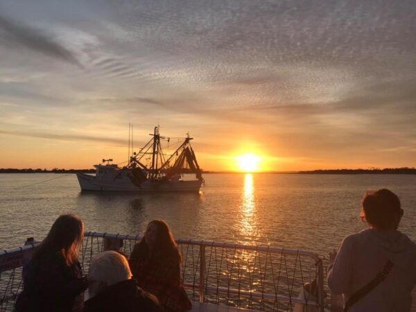 A fishing boat sails on calm water at sunset, with an orange sky and scattered clouds. Four people are in the foreground, silhouetted against the bright sun near the horizon.