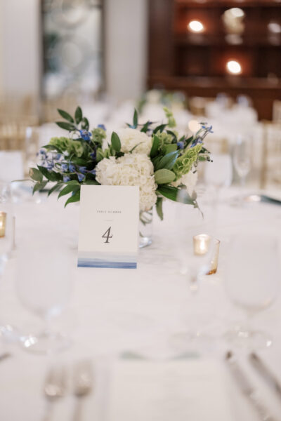 A white table with a floral centerpiece of white and blue flowers, a table number card reading 4, and small gold votive candles, set in an elegant, softly lit event space.