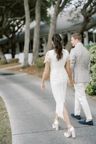 A couple walks hand in hand along a garden path. The woman wears a white lace dress and high heels, while the man wears a light plaid suit and dark shoes. Trees and a building with large windows are in the background.