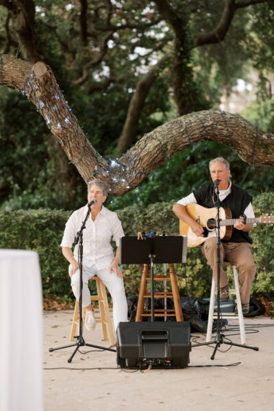 Two musicians perform outdoors beneath a tree with string lights; one sings into a microphone while the other plays an acoustic guitar and sings. Both are seated on stools with music stands and equipment in front of lush greenery.