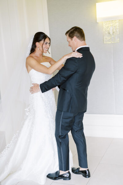 A bride and groom smile and hold each other, sharing a joyful moment indoors. The bride wears a white gown and veil, and the groom wears a dark suit with black shoes. They stand close, gazing at each other happily.