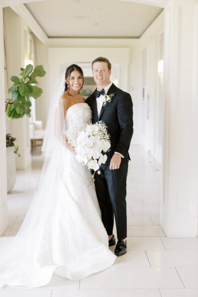 A bride in a white gown and veil stands beside a groom in a black tuxedo. The bride holds a cascading bouquet of white flowers. They smile while posing in a bright, elegant hallway.