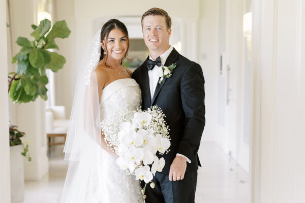 A bride in a white dress and veil stands beside a groom in a black tuxedo, both smiling and holding a bouquet of white flowers in a bright hallway with green plants.