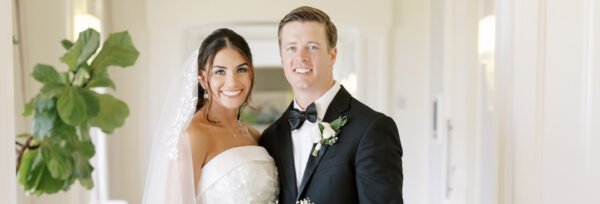A bride in a white wedding dress and veil stands next to a groom in a black tuxedo with a bow tie and boutonniere, both smiling in a bright indoor hallway.