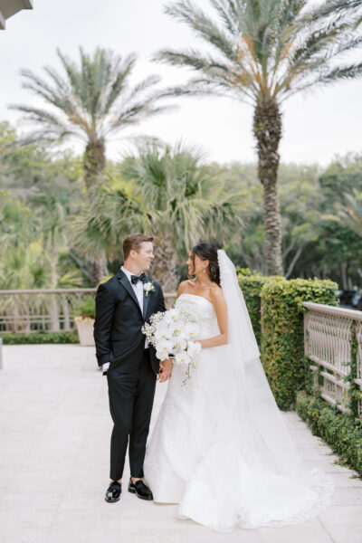 A bride in a white strapless gown and veil holds a bouquet of white flowers, standing next to a groom in a black tuxedo. They gaze at each other outdoors, surrounded by palm trees and greenery.