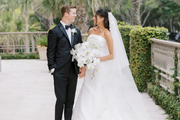 A bride in a white gown and veil holds a bouquet of white flowers and stands smiling with a groom in a black tuxedo. They are outdoors on a stone path, surrounded by green foliage and palm trees.