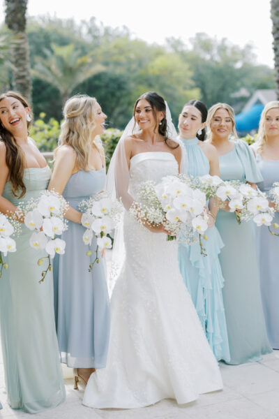 A bride in a white gown stands smiling with five bridesmaids in pale blue dresses, all holding white floral bouquets, outdoors with greenery in the background.