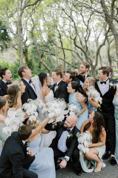 A joyful wedding party poses outdoors, with the bride and groom at the center, surrounded by bridesmaids in light blue dresses holding white bouquets, and groomsmen in black tuxedos, all smiling and celebrating.