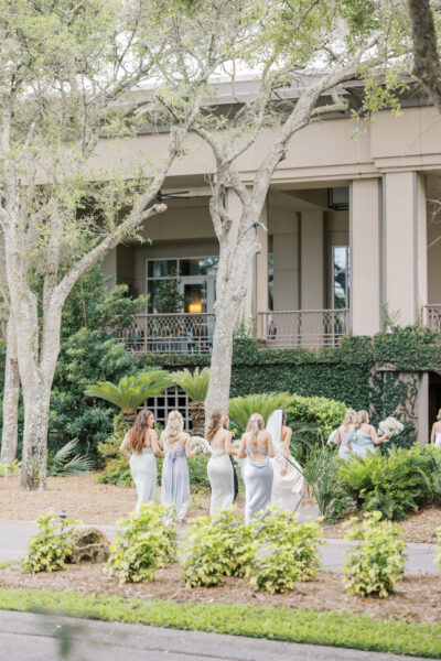 A group of women in light blue dresses walk together outside a building surrounded by trees and greenery on a bright day. The group appears to be celebrating, possibly at a wedding.