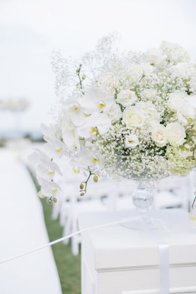 A large floral arrangement in a glass vase featuring white orchids, roses, and babys breath sits atop a pedestal at an outdoor wedding ceremony, with white chairs and an aisle in the background.