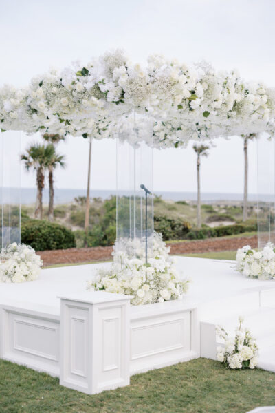 An elegant outdoor wedding altar features white floral arrangements and a canopy of white flowers, set against a backdrop of palm trees and ocean views under a bright sky.