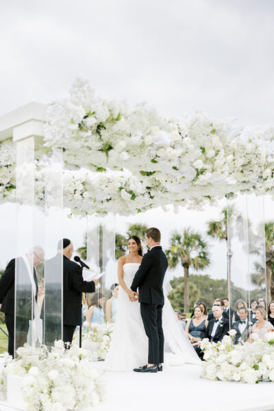 A bride and groom stand under a canopy decorated with white flowers during an outdoor wedding ceremony. Guests are seated around them, and palm trees are visible in the background.