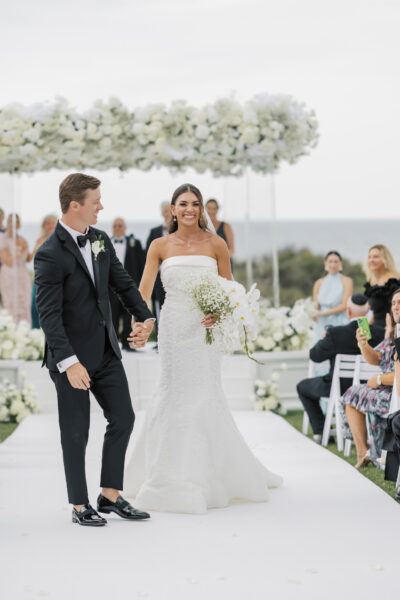 A bride in a strapless white gown and groom in a black tuxedo walk hand in hand down an aisle, smiling, with white floral decorations and guests seated on either side.