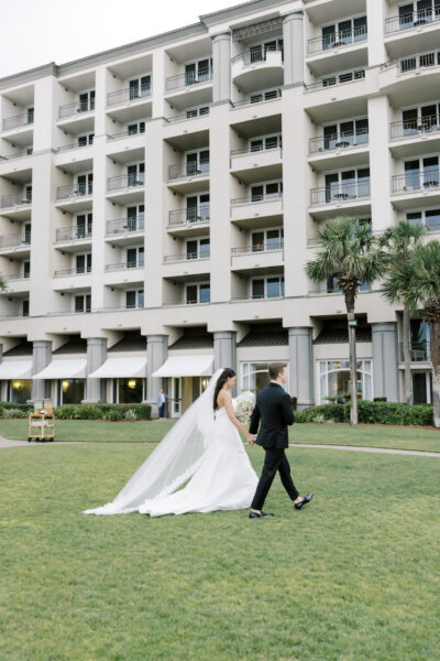 A bride in a long white dress and veil walks hand-in-hand with a groom in a black suit across a green lawn, with a multi-story building and palm trees in the background.