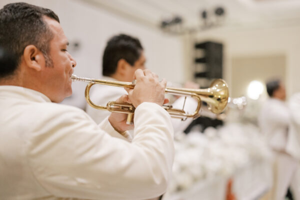 A man in a white suit plays a trumpet at an indoor event, with another musician in the background and white decorations visible. The scene appears festive and elegant.