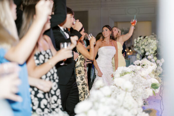 A bride in a strapless white dress stands smiling among guests dressed in formal attire, some raising glasses in a toast, surrounded by white floral arrangements at a celebration.