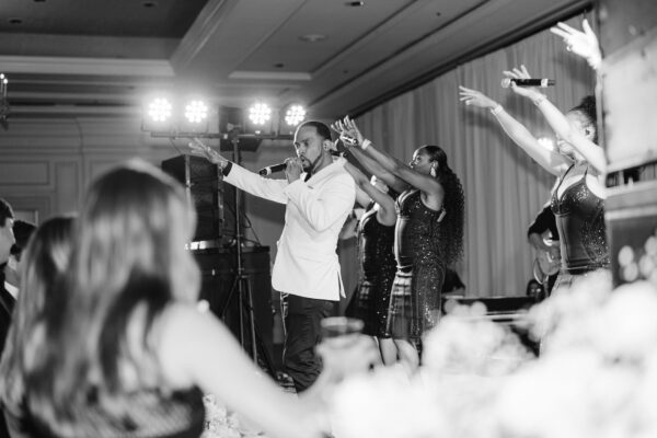 A male singer in a white jacket performs on stage with three female backup dancers, all with arms raised, in front of an audience at an indoor event. The scene is captured in black and white.