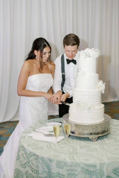A bride and groom, both smiling, cut a tall white wedding cake decorated with flowers. They stand together at a table with two champagne glasses and white napkins on a green-patterned tablecloth.