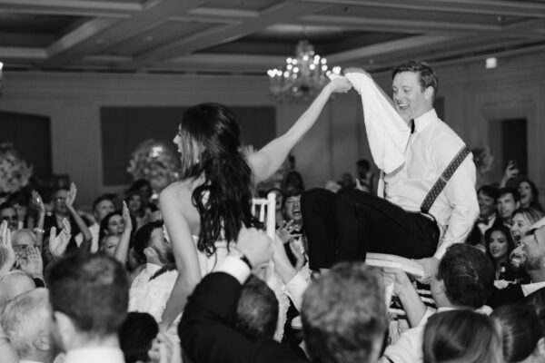A joyful couple is lifted on chairs by a crowd at a lively wedding reception, holding hands and smiling as guests cheer around them in a grand ballroom.