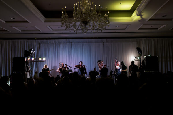 A band performs on a brightly lit stage in front of a crowd in a ballroom with a chandelier and draped white curtains. The audience is mostly in shadow.