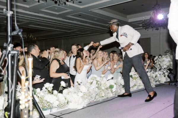 A performer in a white jacket and hat fist-bumps an excited audience at an indoor event, with white flowers lining the stage and people smiling, cheering, and taking photos.