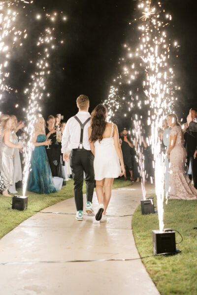 A couple walks arm in arm down a path lined with sparklers, surrounded by cheering guests in formal attire at night, celebrating in a festive outdoor setting.