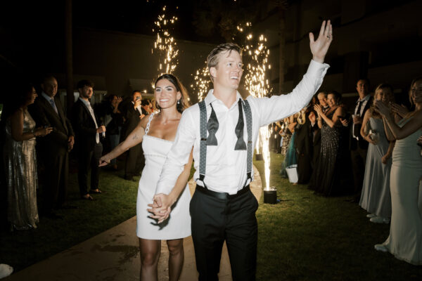 A newlywed couple, smiling and holding hands, walks through a crowd of cheering guests at night. Sparklers light up the background as the groom waves and the bride beams with joy. The guests are dressed in formal attire.