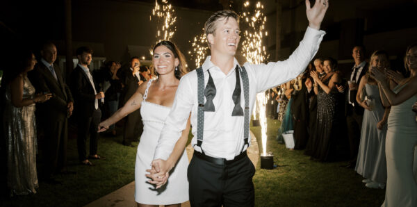 A bride in a short white dress and a groom in a white shirt with suspenders smile and hold hands, walking past guests and sparklers at an outdoor nighttime celebration.