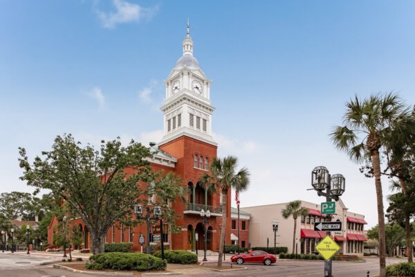A historic red-brick courthouse with a white clock tower stands on a corner surrounded by palm trees, street signs, and a red car parked nearby under a clear blue sky.