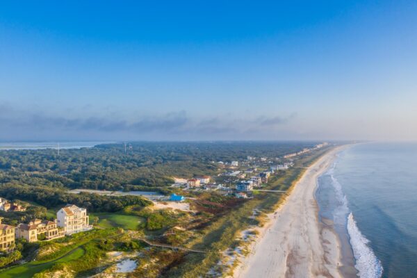 Aerial view of a long sandy beach bordered by ocean on one side and coastal houses and lush greenery on the other, under a clear blue sky.