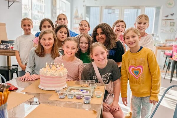 A group of smiling kids gather around a table with a pink and white birthday cake topped with lit candles. Art supplies are on the table, and the setting is a bright, cheerful room.