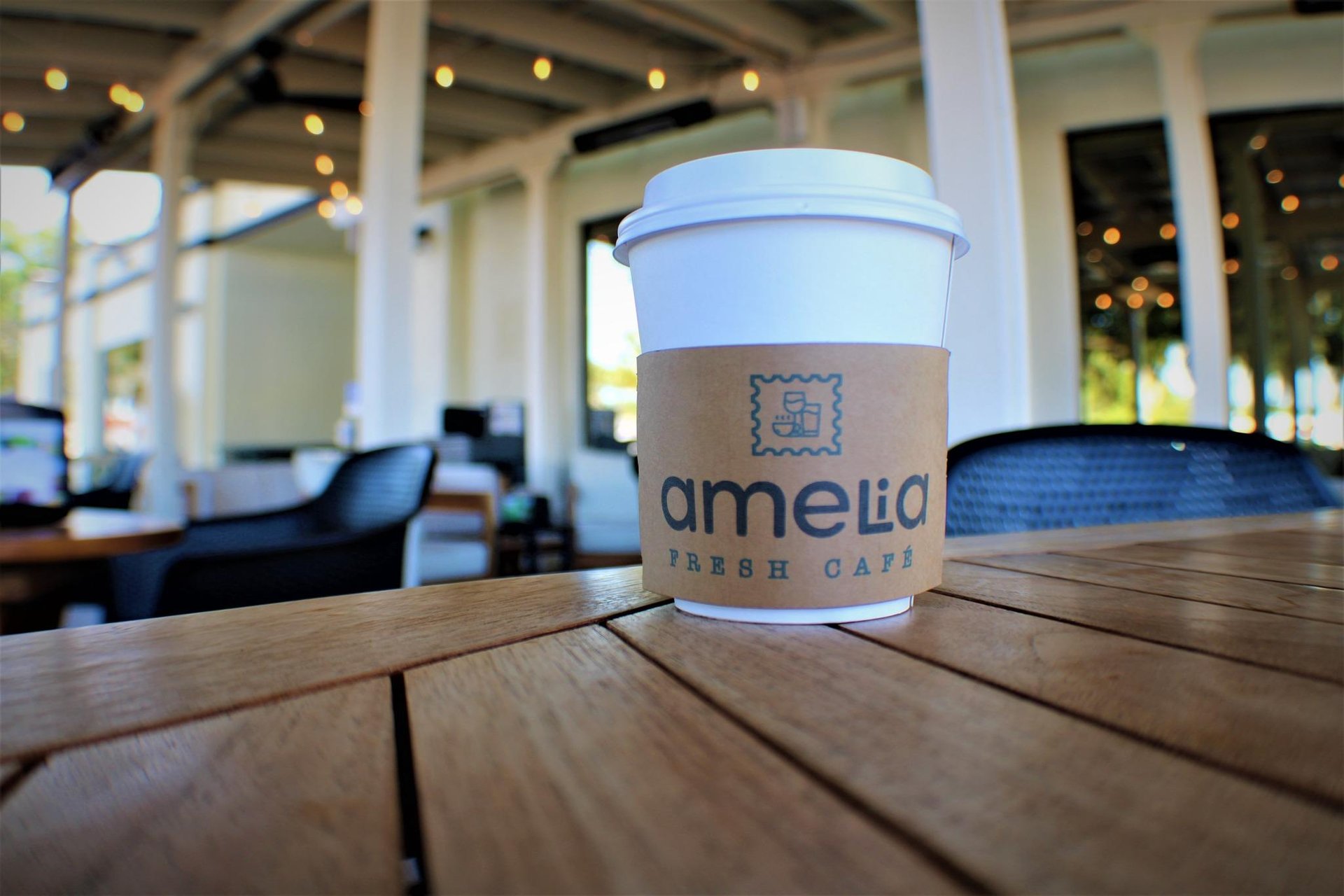 A white takeaway coffee cup with a cardboard sleeve reading amelia FRESH CAFE sits on a wooden table in an outdoor café with string lights and lounge chairs in the background.